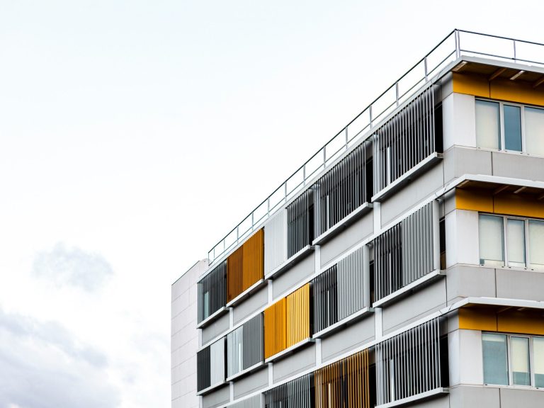 Edificio moderno con fachada gris y paneles amarillos y anaranjados. Cielo nublado.